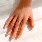 A close-up of a hand wearing a delicate amber ring with a small amber-colored stone and thin silver-toned metal bands, set against a bright white background with soft shadows.