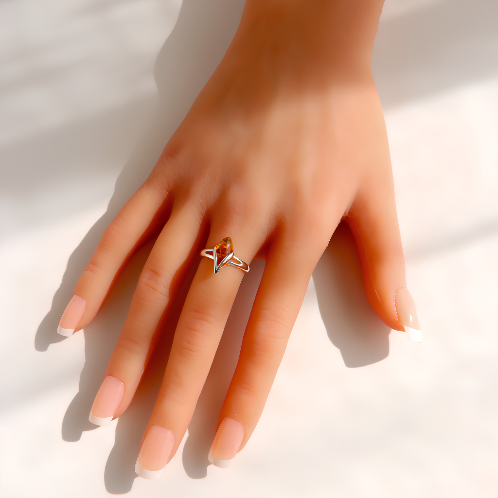 A close-up of a hand wearing a delicate amber ring with a small amber-colored stone and thin silver-toned metal bands, set against a bright white background with soft shadows.