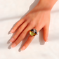 A hand with a French manicure wearing a large silver amber ring featuring a round, marbled green and yellow stone with a small silver frog motif on top, set against a bright white background with soft shadows.