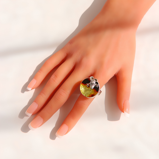 A hand with a French manicure wearing a large silver amber ring featuring a round, marbled green and yellow stone with a small silver frog motif on top, set against a bright white background with soft shadows.