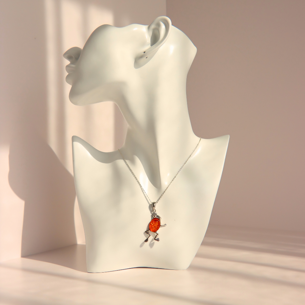 A silver amber frog pendant necklace with a small frog displayed on a white female mannequin bust against a warm beige background.