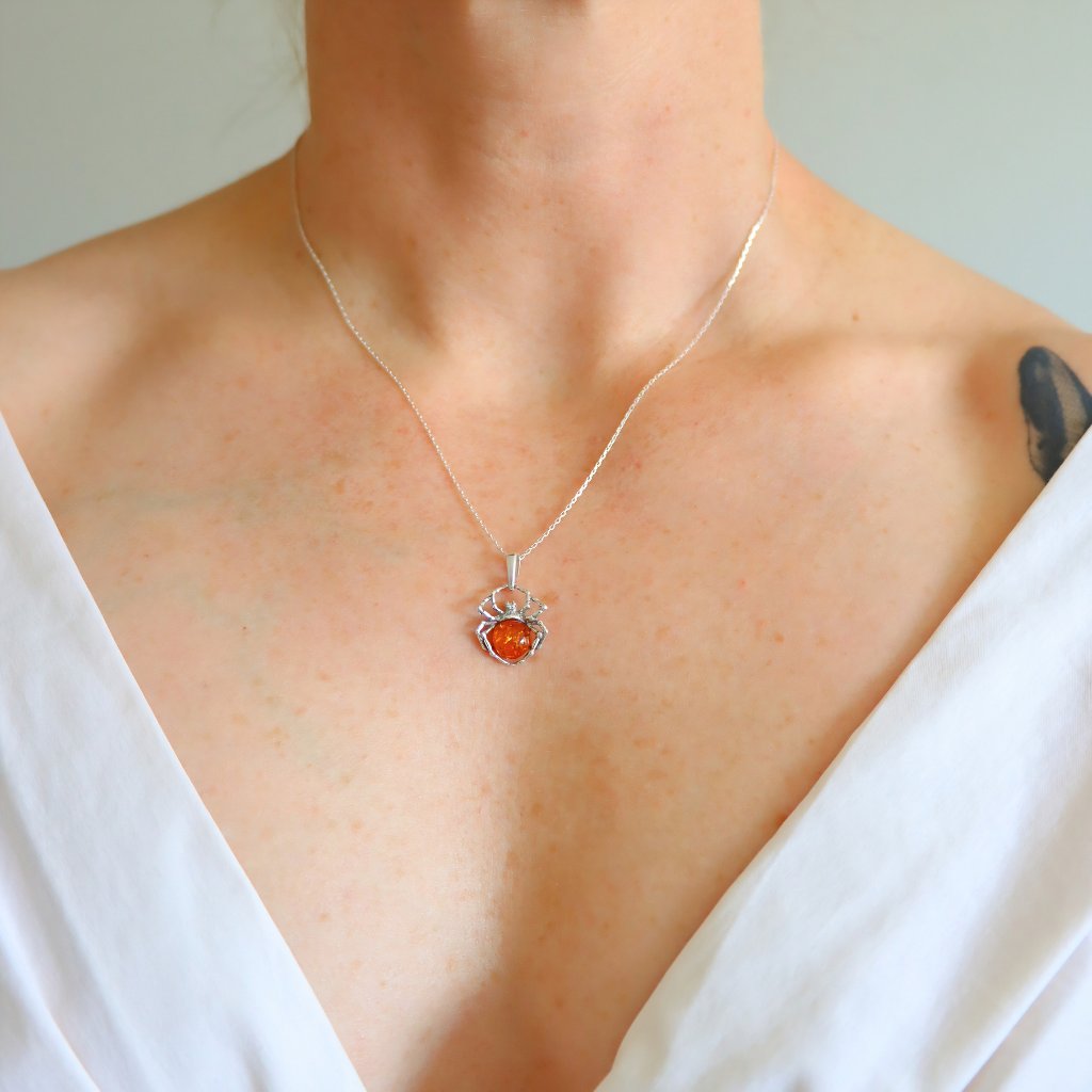 A close-up of a person's neck and chest wearing a delicate silver chain necklace with a small amber spider pendant featuring a vibrant orange center.