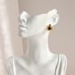 A gold-toned spider-shaped stud earring with a dark red center, displayed on a glossy white female mannequin bust against a soft beige background.