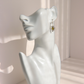 A silver-toned heart-shaped green amber drop earring with a green center stone, shown hanging on a white ceramic female bust against a softly lit beige background.
