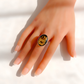 A amber cameo ring with a black oval face featuring a raised floral and fairy motif, worn on a hand with a French manicure against a bright white background.