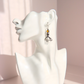 A silver-toned dangle amber octopus earring with a small amber-orange bead and silver tentacles shown on a white female bust against a soft pink studio background.