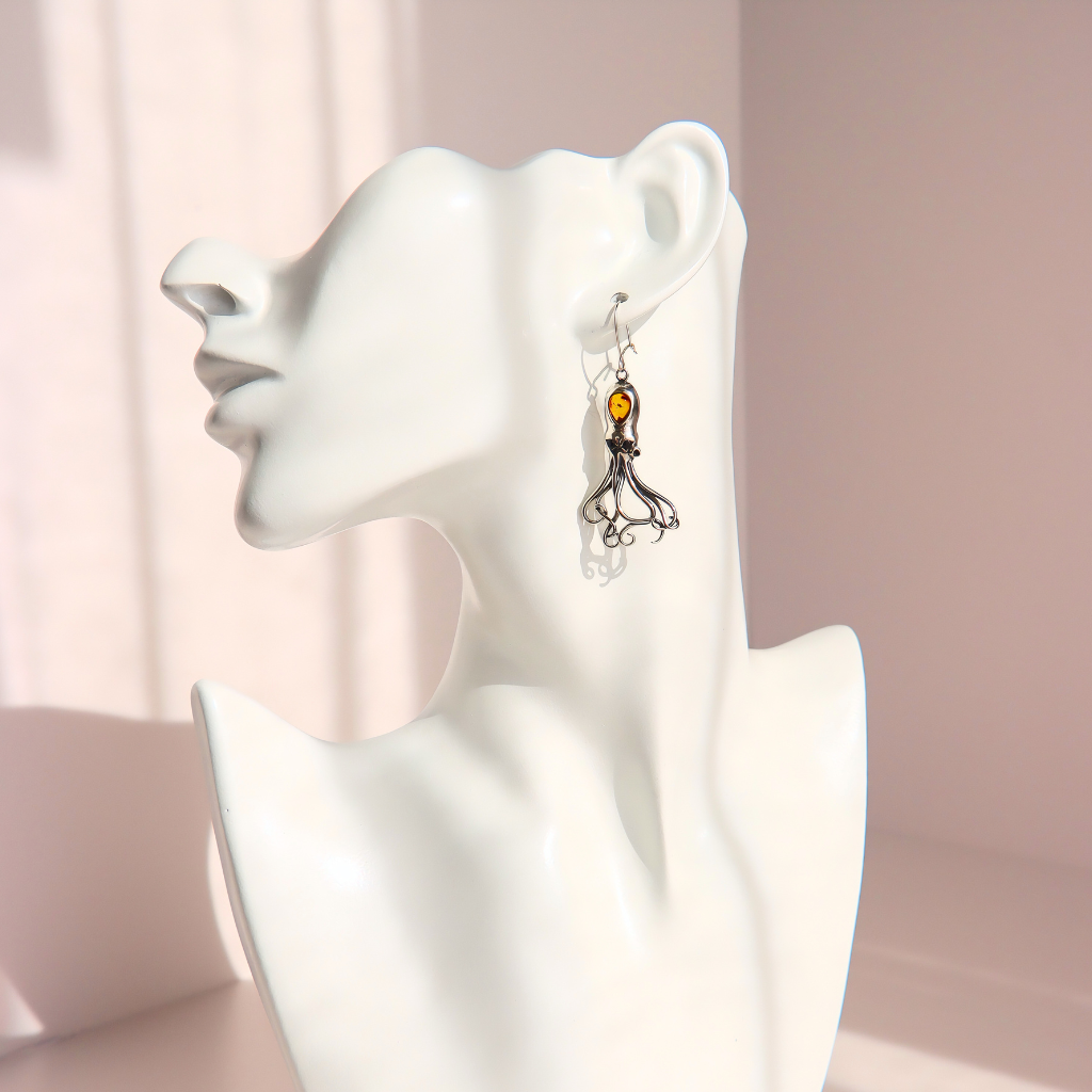 A silver-toned dangle amber octopus earring with a small amber-orange bead and silver tentacles shown on a white female bust against a soft pink studio background.