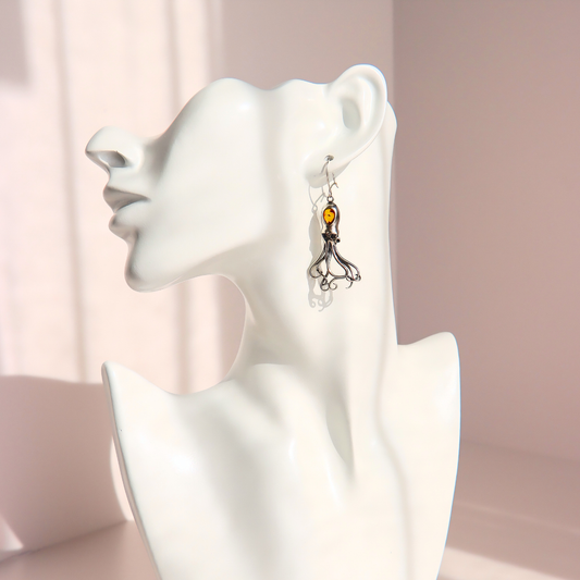 A silver-toned dangle amber octopus earring with a small amber-orange bead and silver tentacles shown on a white female bust against a soft pink studio background.