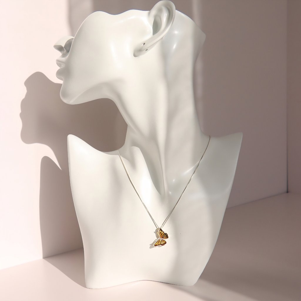 A delicate silver amber necklace with a butterfly pendant, displayed on a white ceramic female bust mannequin against a soft pink background.