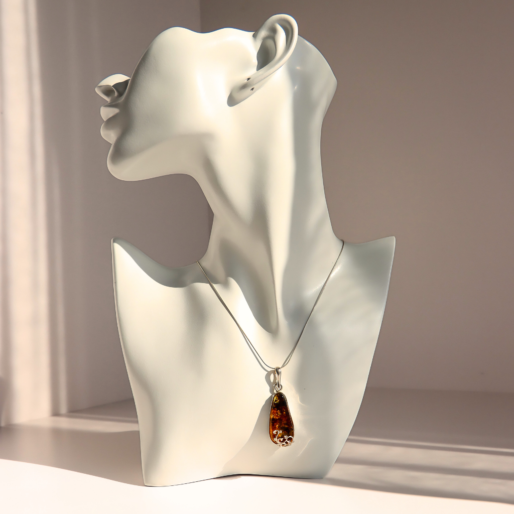 A silver-toned teardrop green amber pendant necklace with a brown amber-like drop, displayed on a glossy white female bust mannequin against a warm beige background.