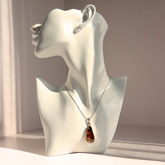 A silver-toned teardrop green amber pendant necklace with a brown amber-like drop, displayed on a glossy white female bust mannequin against a warm beige background.