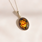 An oval pendant amber necklace featuring a yellow and orange scarab design on a dark background, suspended from a silver chain against a light beige backdrop.