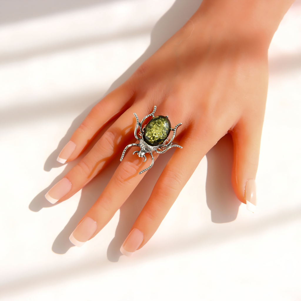 A hand with French manicure wearing a silver green amber beetle ring with a large, textured green oval gemstone, set against a bright white background with soft diagonal shadows.