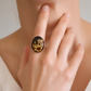 A close-up lifestyle shot of a person wearing a large oval amber cameo ring on their finger. The ring features a black enamel background with a detailed gold floral and butterfly design.
