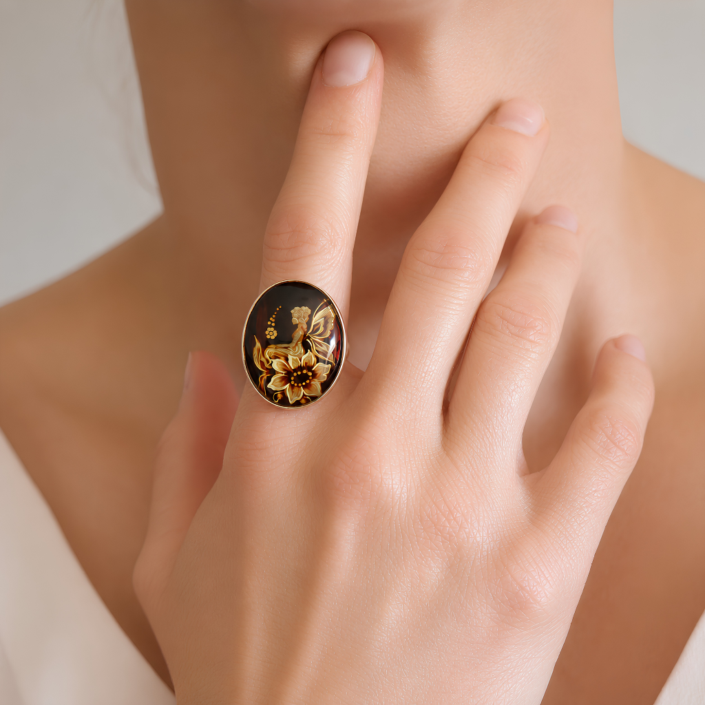 A close-up lifestyle shot of a person wearing a large oval amber cameo ring on their finger. The ring features a black enamel background with a detailed gold floral and butterfly design.