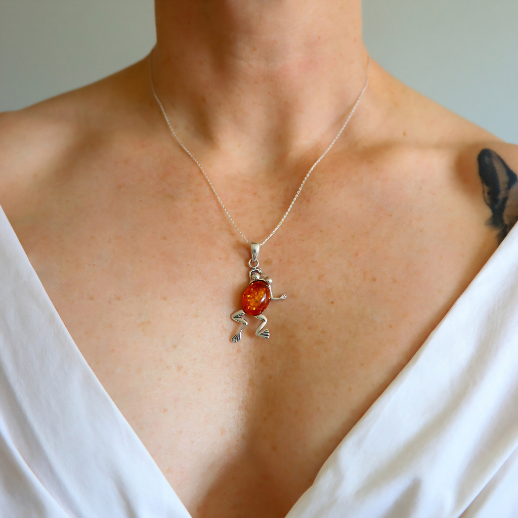 A close-up of a person wearing a delicate silver necklace with a small, silver frog pendant featuring a round amber-orange center, shown against the wearer's chest.