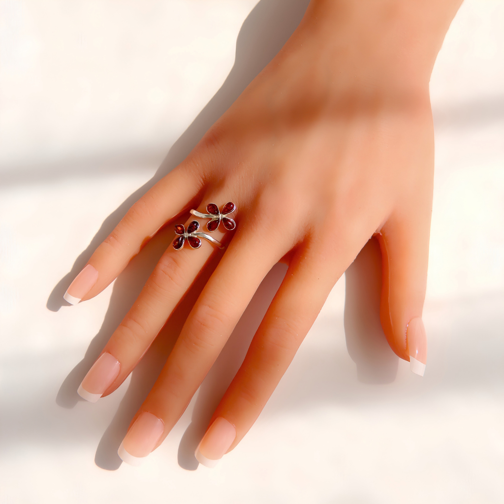 A hand with a French manicure wearing a silver Baltic amber flower ring decorated with Two small dark red flower-shaped charms, set against a bright white background with soft shadows.