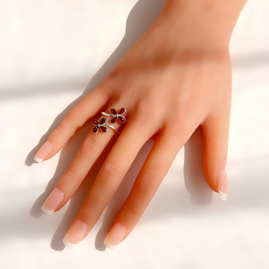 A hand with a French manicure wearing a silver Baltic amber flower ring decorated with Two small dark red flower-shaped charms, set against a bright white background with soft shadows.