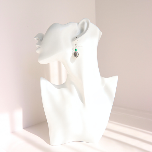 A silver-toned western malachite dangle earring with a round dark charm, displayed on the ear of a white ceramic mannequin bust against a soft beige background.