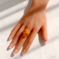 A gold amber ring with an oval amber cabochon gemstone, worn on a hand with a French manicure, set against a bright white background with soft diagonal shadows.