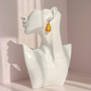 A white ceramic female bust display wearing a single Baltic amber dangle earring with a silver hoop, photographed in warm sunlight against a pale pink wall.
