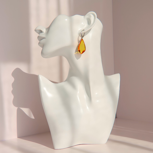 A white ceramic female bust display wearing a single Baltic amber dangle earring with a silver hoop, photographed in warm sunlight against a pale pink wall.