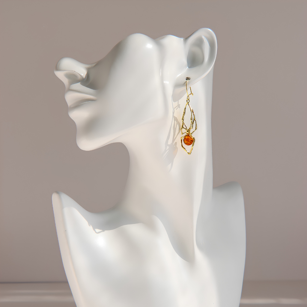 A pair of gold-toned dangle amber spider earrings with a small amber-orange bead, displayed on a white ceramic female bust against a soft beige background.