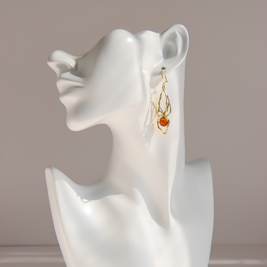 A pair of gold-toned dangle amber spider earrings with a small amber-orange bead, displayed on a white ceramic female bust against a soft beige background.