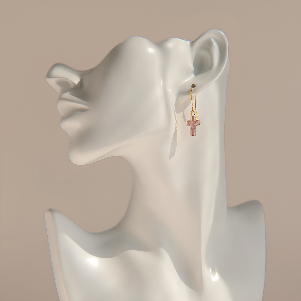 A pair of small gold rose quartz cross drop earrings displayed on a white ceramic female bust against a warm beige background.