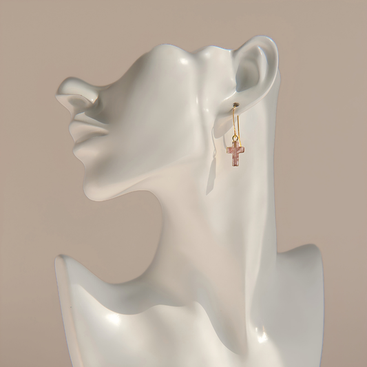 A pair of small gold rose quartz cross drop earrings displayed on a white ceramic female bust against a warm beige background.