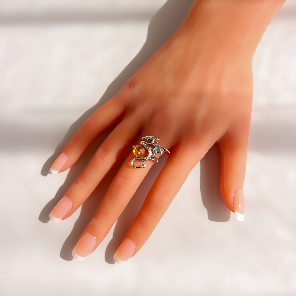 A hand with a French manicure wearing a silver amber dragon ring with a small orange gemstone, set against a bright white background with soft shadows.