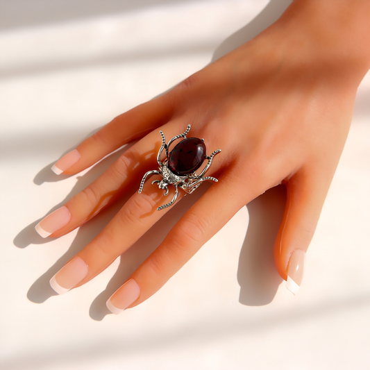 A hand wearing a silver scarab ring with a large oval dark red gemstone, set against a bright white background with soft shadows.