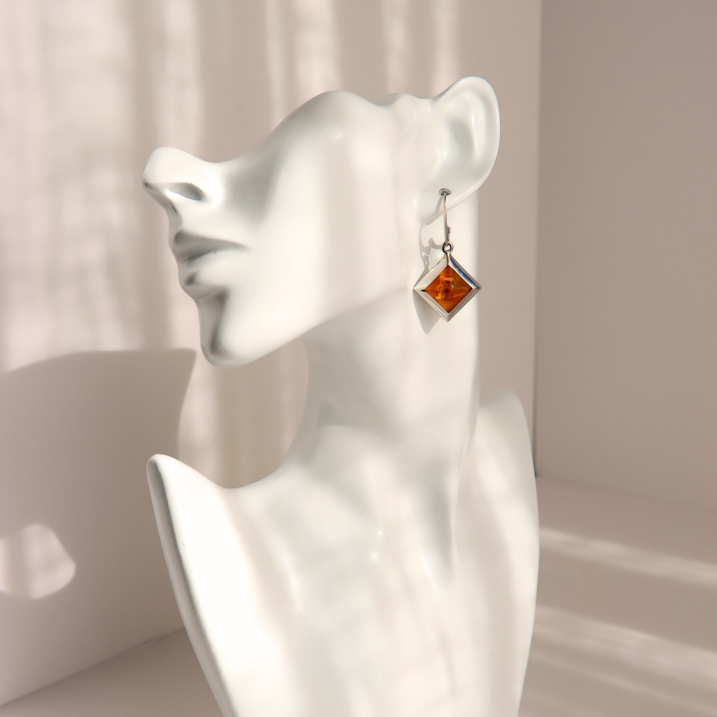 A single silver-toned dangle amber earring with a square amber-orange stone, displayed on a white ceramic female bust against a softly lit beige background.