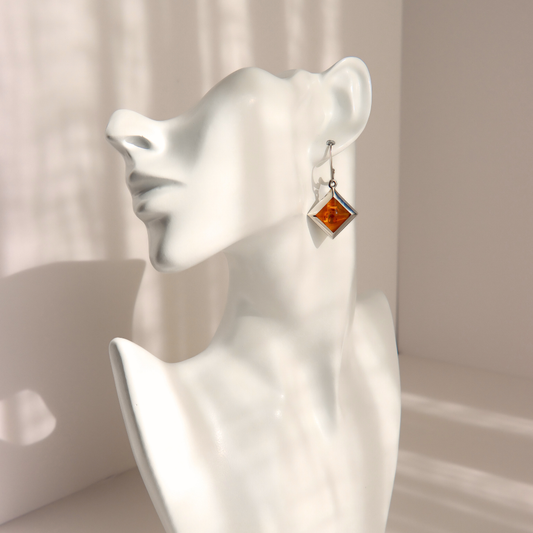 A single silver-toned dangle amber earring with a square amber-orange stone, displayed on a white ceramic female bust against a softly lit beige background.