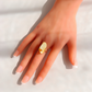 A hand with a French manicure wearing a white amber gold ring with a large, irregularly shaped cream-colored stone, set against a bright, softly shadowed white background.