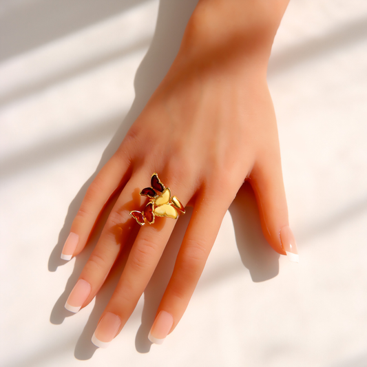 A gold amber butterfly ring worn on a hand with a French manicure, set against a bright white background with soft diagonal shadows.