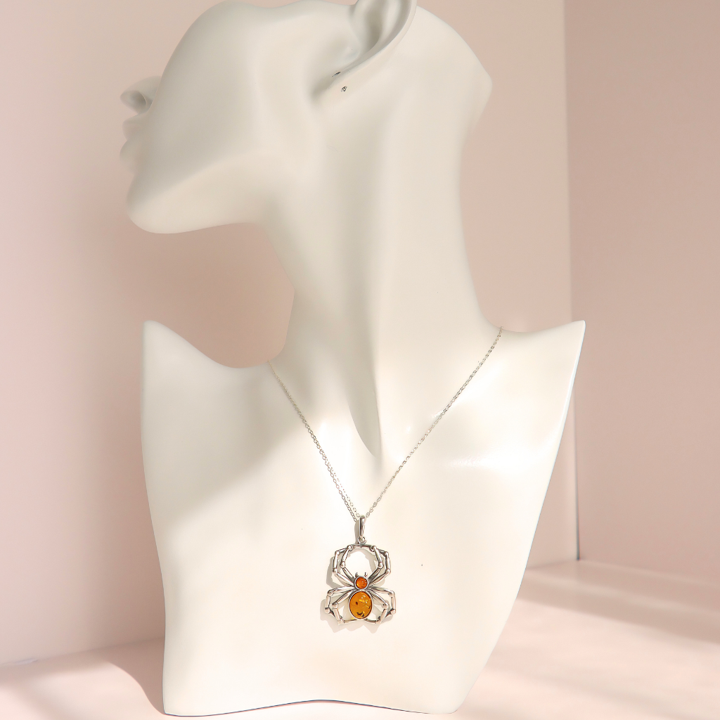 A silver spider pendant amber necklace with a central amber-orange gemstone, displayed on a white female mannequin bust against a soft pink background.