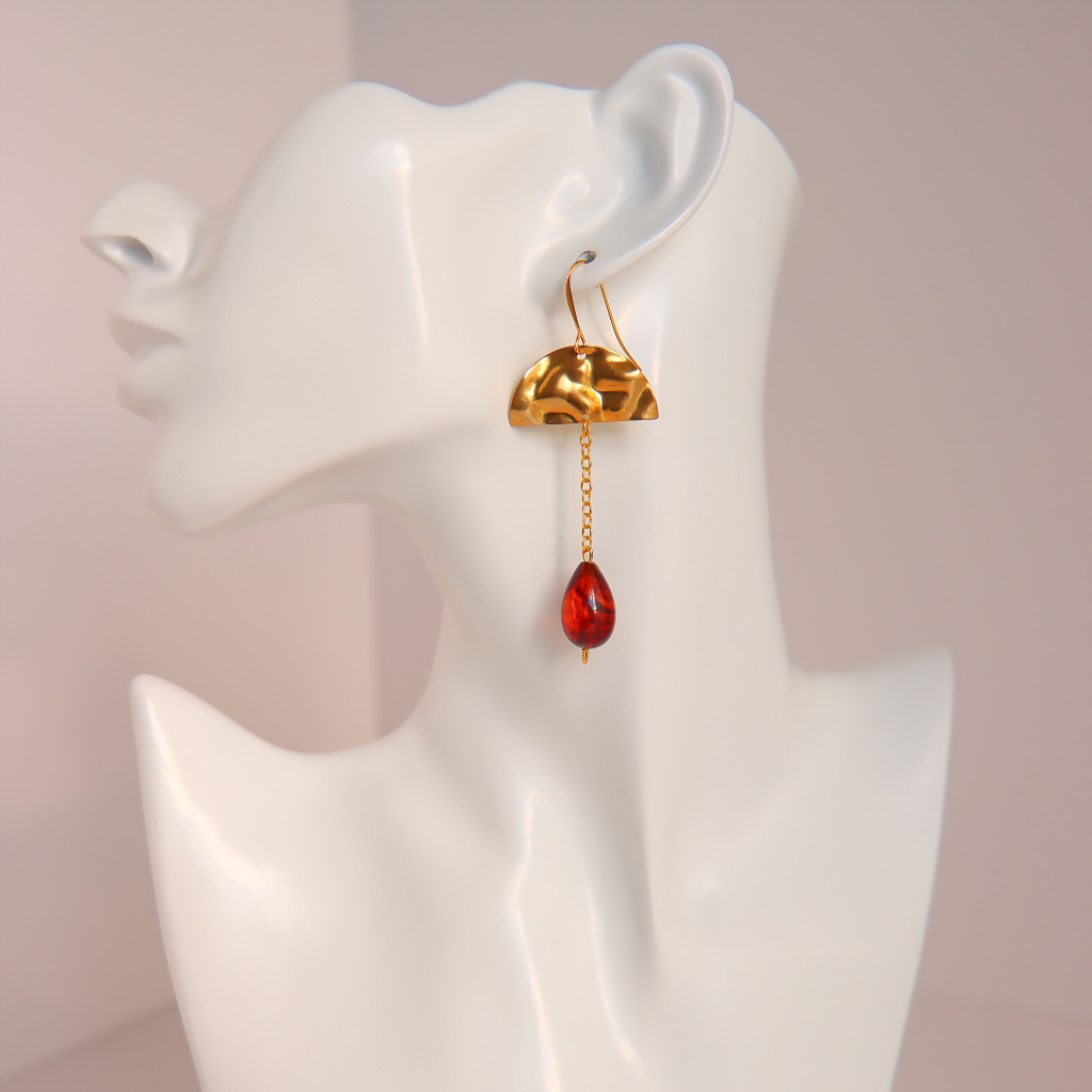A gold dangle vintage Lucite amber earring with a textured dome-shaped top and a dangling red Lucite teardrop bead, displayed on a white ceramic mannequin head against a soft pink background.