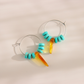 A pair of silver & turquoise hoop earrings with turquoise blue beads and amber-colored drop charms, displayed on a warm beige background with soft shadows.