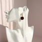 A single dark amber rose earring with a gold hoop and hook, displayed on the ear of a white ceramic female bust against a soft pink background.