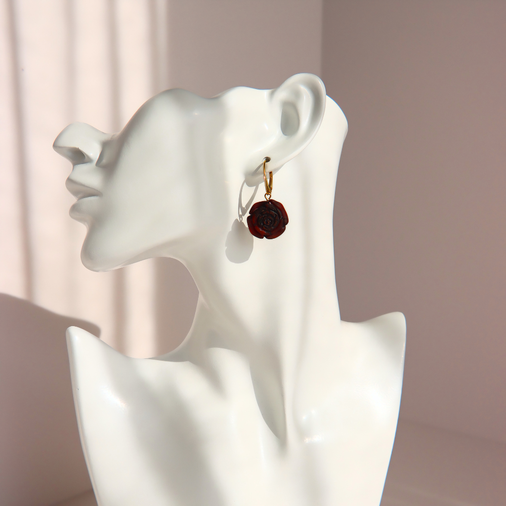 A single dark amber rose earring with a gold hoop and hook, displayed on the ear of a white ceramic female bust against a soft pink background.