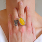 A close-up lifestyle shot of a person's hand wearing a large sterling silver amber poison ring with a smooth, oval yellow cabochon gemstone.