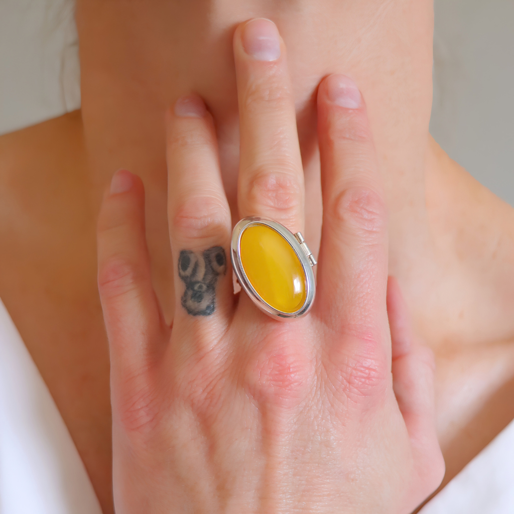 A close-up lifestyle shot of a person's hand wearing a large sterling silver amber poison ring with a smooth, oval yellow cabochon gemstone.