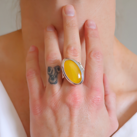 A close-up lifestyle shot of a person's hand wearing a large sterling silver amber poison ring with a smooth, oval yellow cabochon gemstone.