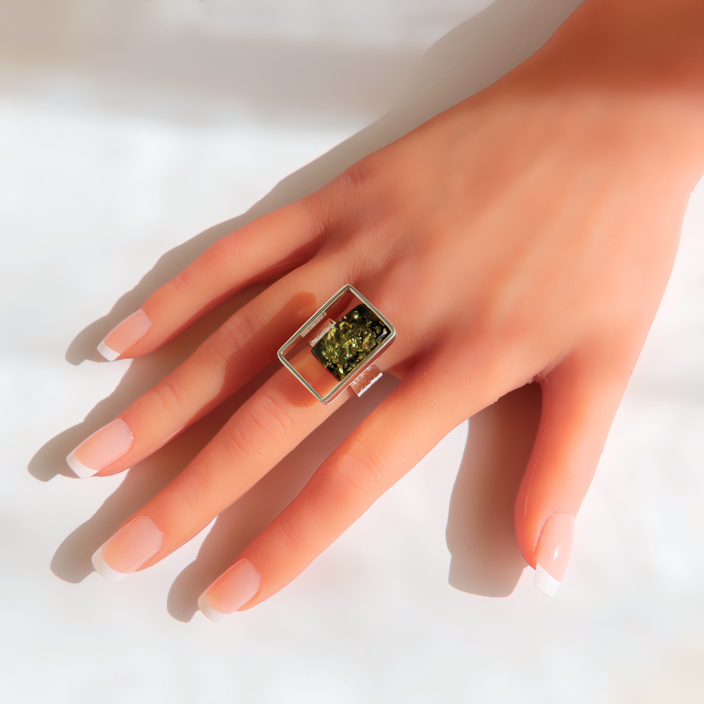 A silver-toned amber ring with a rectangular green gemstone, worn on a hand with a French manicure against a bright white background.