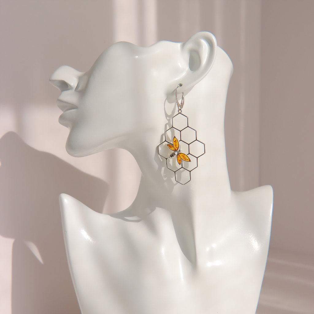 A silver-toned honeycomb dangle earring with two small yellow bee charms, displayed on a white ceramic female bust against a soft beige background.