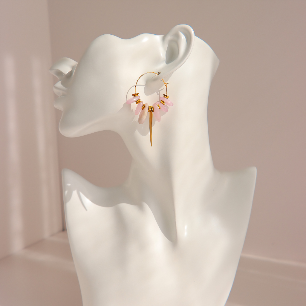 A pair of gold kunzite hoop earrings with dangling pink beads and gold charms, displayed on a white ceramic mannequin head against a soft pink background.