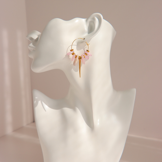 A pair of gold kunzite hoop earrings with dangling pink beads and gold charms, displayed on a white ceramic mannequin head against a soft pink background.