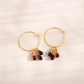 Gold hoop earrings with three dangling teardrop beads in a deep burgundy red, set against a warm off-white background with soft diagonal shadows.