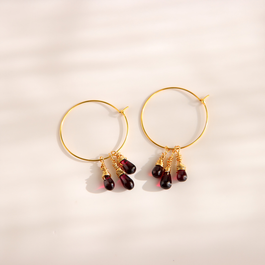 Gold hoop earrings with three dangling teardrop beads in a deep burgundy red, set against a warm off-white background with soft diagonal shadows.
